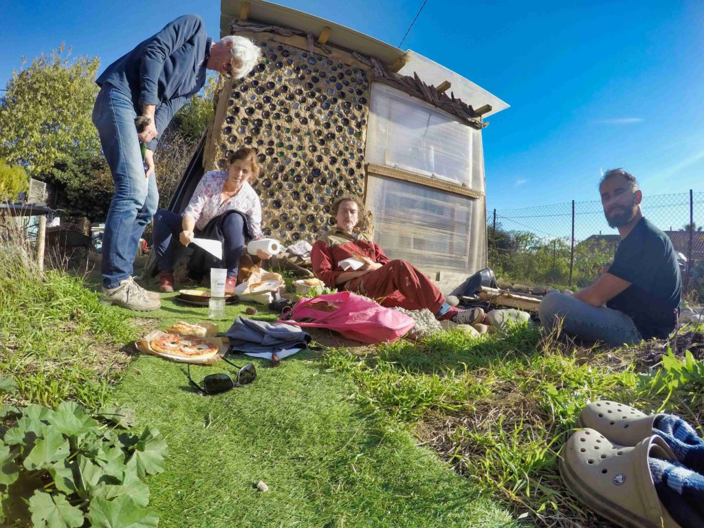 des gens savourant une pause déjeuner devant une serre bioclimatique sous un beau ciel bleu et ensoleillé, dans l'herbe au pied d'un mur en bouteilles de verre.